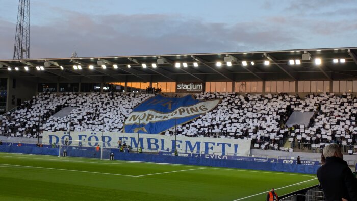 Publikhav på en fotbollsarena med ett stort tifo som visar IFK Norrköpings emblem och texten "DÖ FÖR EMBLEMET", åskådare håller upp vita papper.