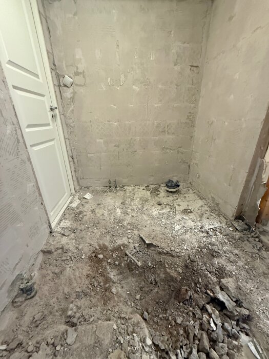 Rivet golv med exponerad golvbrunn i ett rum under renovering, smutsigt och ojämt golv samt vit dörr i bakgrunden.