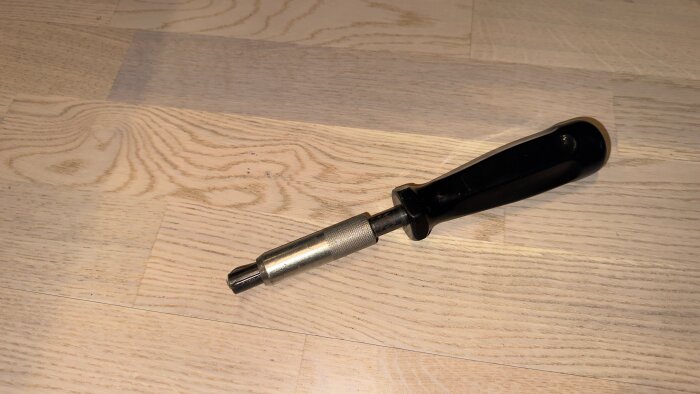 Verktyg med svart handtag och metallspets, eventuellt för säkerhetsfattning vid belysningsstolpar, liggande på ett trägolv.