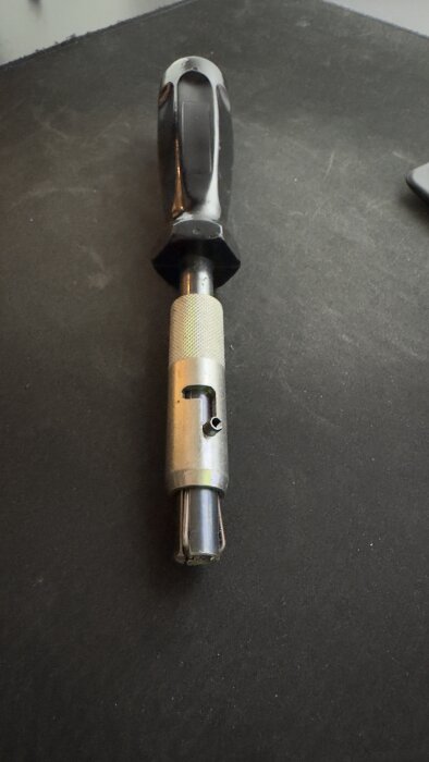 Verktyg med svart handtag och metallklämma, möjligtvis för säkerhetsfattning på belysningsstolpe, placeras på en mörk yta.