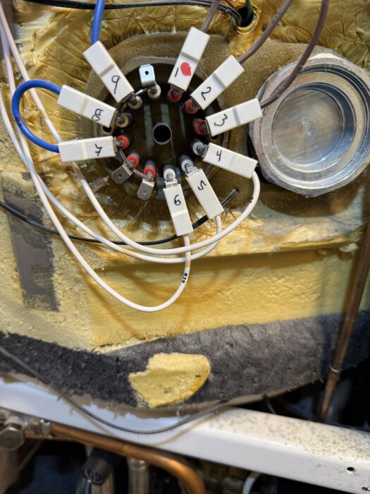 Kopplingspanel på elpatronen med numrerade anslutningar och kablar på en värmepump, omgiven av isolering.