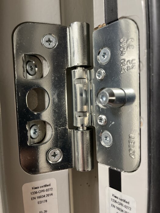 Närbild av gångjärnet på en branddörr, med skruvar i olika färger för justering. En etikett med Kiwa-certifiering syns också.
