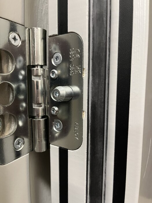 Närbild på gångjärnet av en branddörr med synliga skruvar och märkningar, vilket visar var justering behövs för att korrigera dörrens hängning.