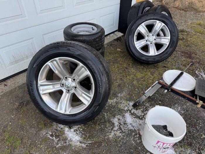 Fyra däck med aluminiumfälgar ligger på marken vid en garageport, samt en hink och verktyg i närheten.