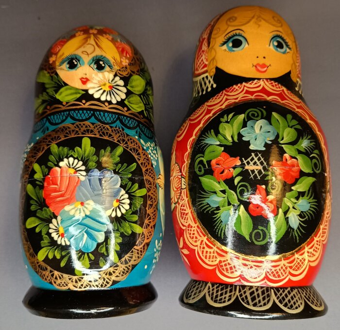 Två matryoshkadockor med färgglada blommönster, en i blå och en i röd dräkt, placerade mot en grå bakgrund.