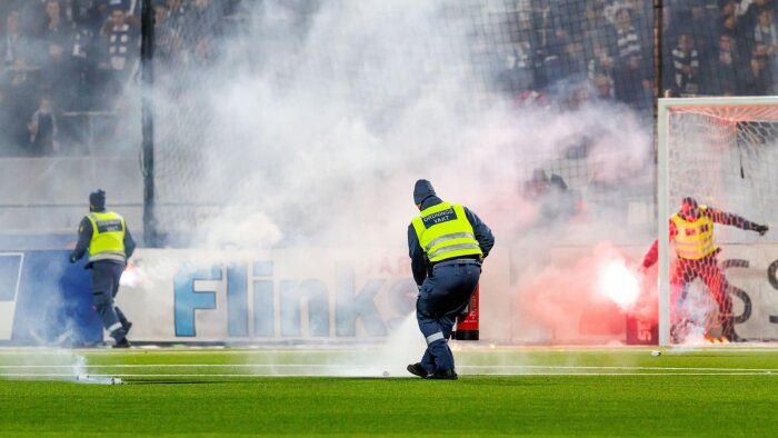 Ordningsvakter på fotbollsplan släcker brinnande rökfacklor, inramat av tjock vit rök.