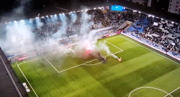 Fotbollsstadion med rök och eldar på planen, delvis tomma läktare syns i bakgrunden.