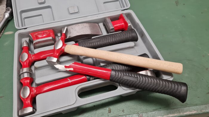 En karosserihammare med ett nytt trähandtag jämte trasiga hammare med röda handtag i en verktygslåda.