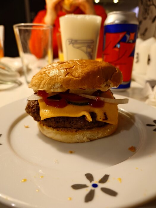 Smashad burgare med ost, lök och ketchup på en tallrik, omgiven av drycker och en person i bakgrunden.