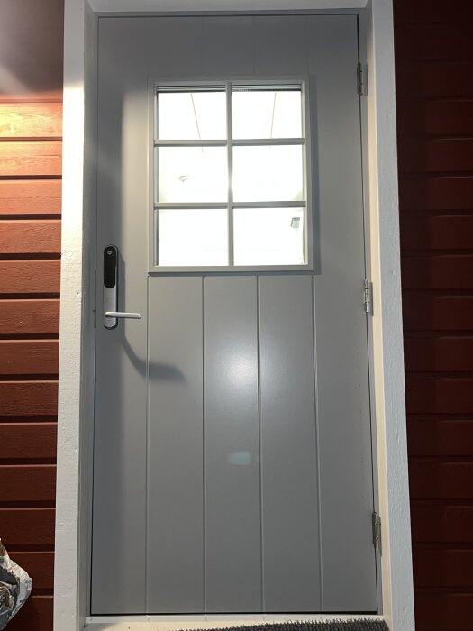 Grå ytterdörr med glasruta i fyrdelat mönster, bredvid röd träpanel. Dörrhandtag och elektroniskt lås på vänster sida.