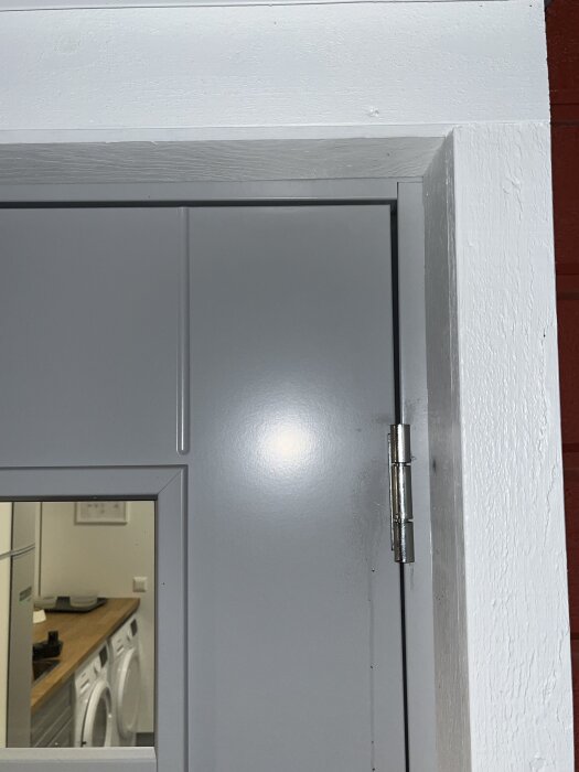 Grå dörr med litet fönster och synlig gångjärn, interiör med tvättmaskiner och träbänkskiva syns genom fönstret.