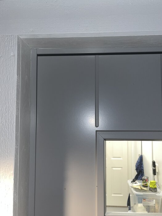 Närbild av en grå dörr med glasruta som visar insidan av ett rum med plastlåda, kläder och verktyg på hyllor.