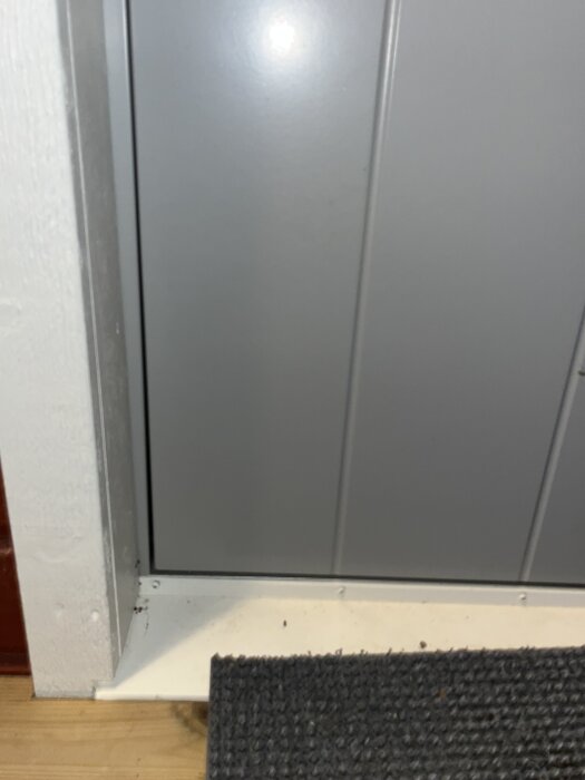Grå dörr med vit karm och mörk dörrmatta framför.