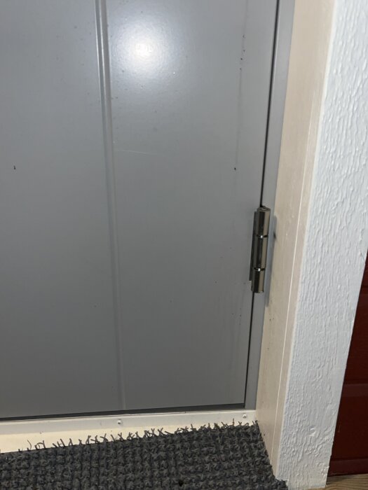 Grå dörr med stånggångjärn och vit dörrkarm, delvis på en mörk dörrmatta.