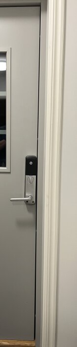 Grå dörr med silverfärgat handtag och digitalt Yale-lås, delvis öppnad, med innervägg och karm i ljus ton.