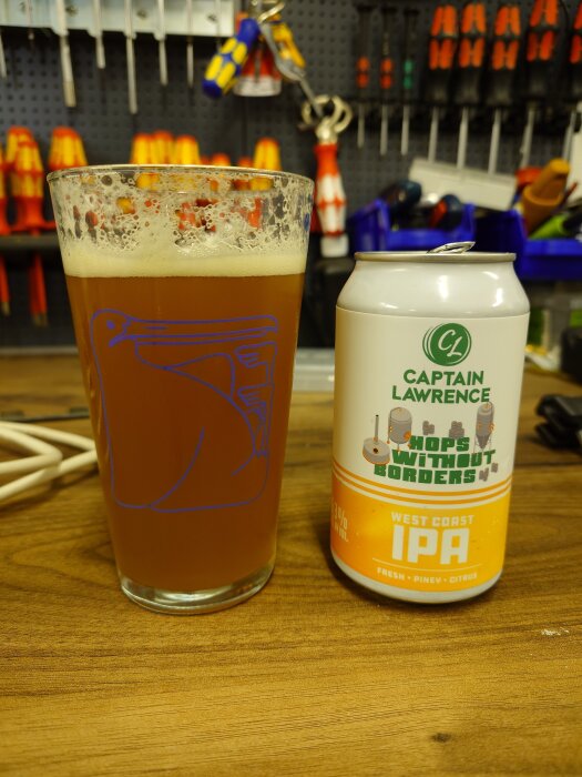 Kapsylöppnad burk med Captain Lawrence IPA bredvid ett glas öl, fotad på en bänkyta med verktygshållare i bakgrunden.