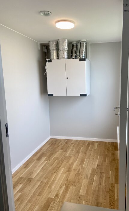 Rum med trägolv och ljusgrå väggar, ventilationsskåp monterat nära taket.