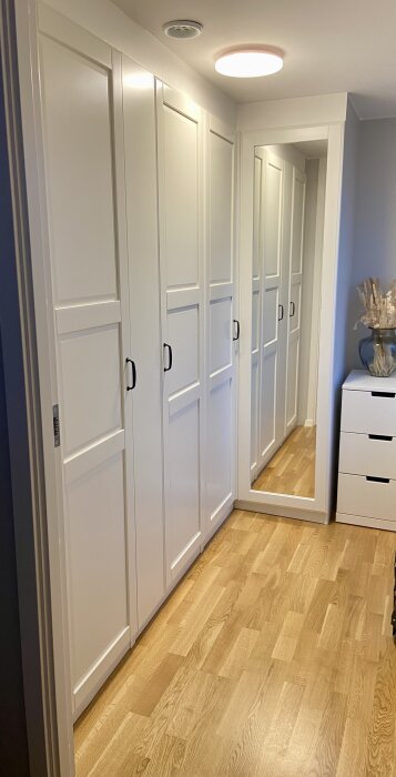 Vit platsbyggd garderob med spegeldörr och svarta handtag, placerad i ett modernt inrett rum med trägolv och ett runt takljus.