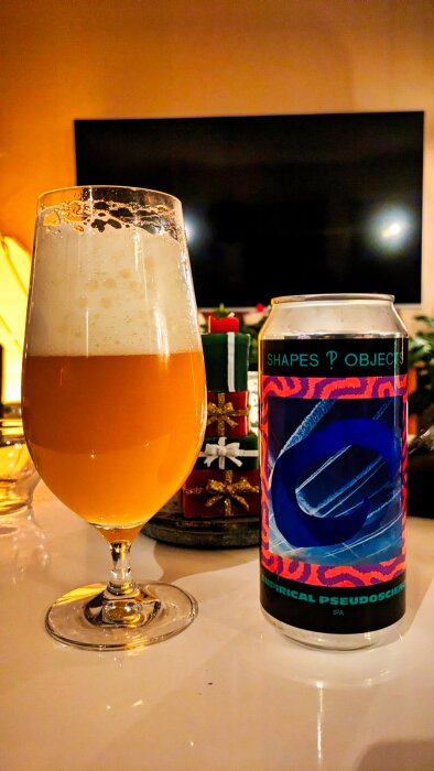 Ett glas skummande öl bredvid en färgglad ölburk på ett bord med levande ljus i bakgrunden.