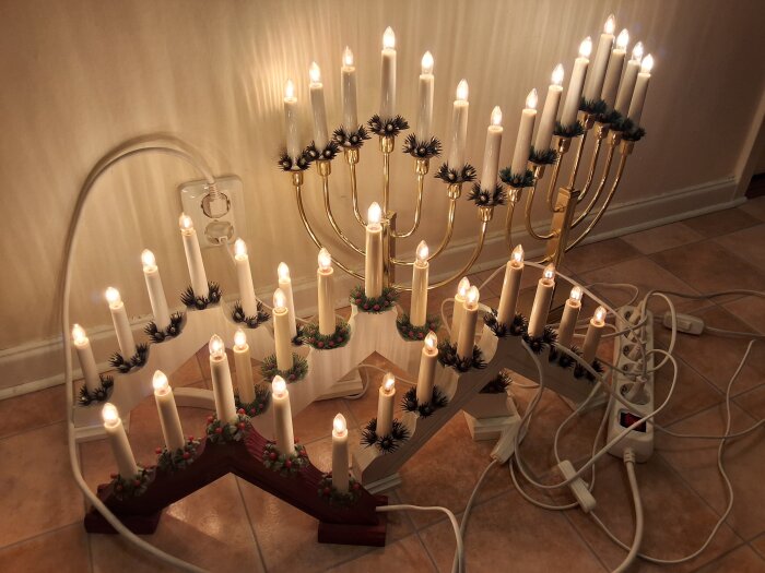 Adventsljusstakar på golv med elektriska ljus tända, kopplade till grenuttag. En femarmad och en sjuarmad ljusstake syns.