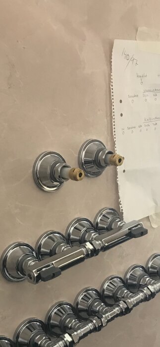 Kromade vattenledningar och ventiler på beige kakelvägg med ett papper med handskrivna anteckningar tejpat bredvid.