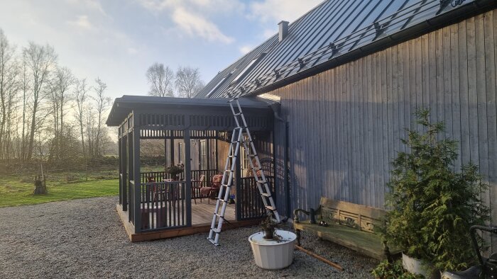 Bild på en utomhusterass intill en grå byggnad med en stege lutad mot taket. Grus på marken och växter i krukor.