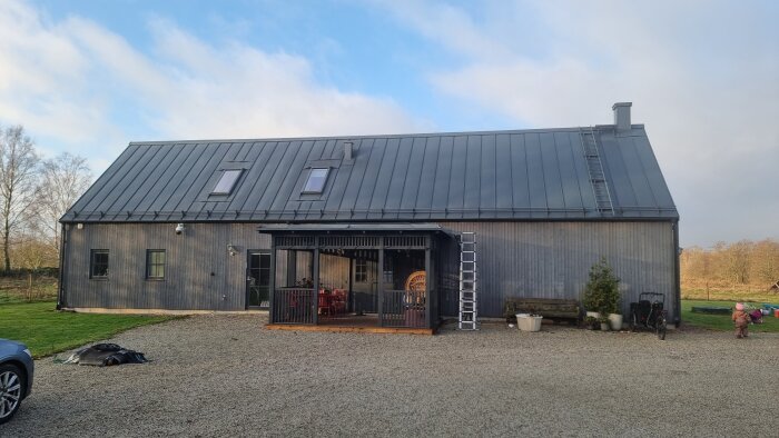 Modernt hus med grått ståltak och träpanel, veranda med bord och stolar, stege längs taket, grusuppfart och omgivande grönska.
