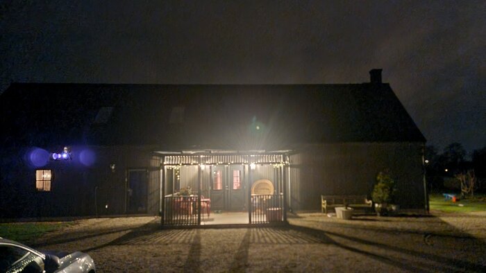 Nattbild av husfasad med belysning över veranda och dekorationer. Mörk himmel i bakgrunden.