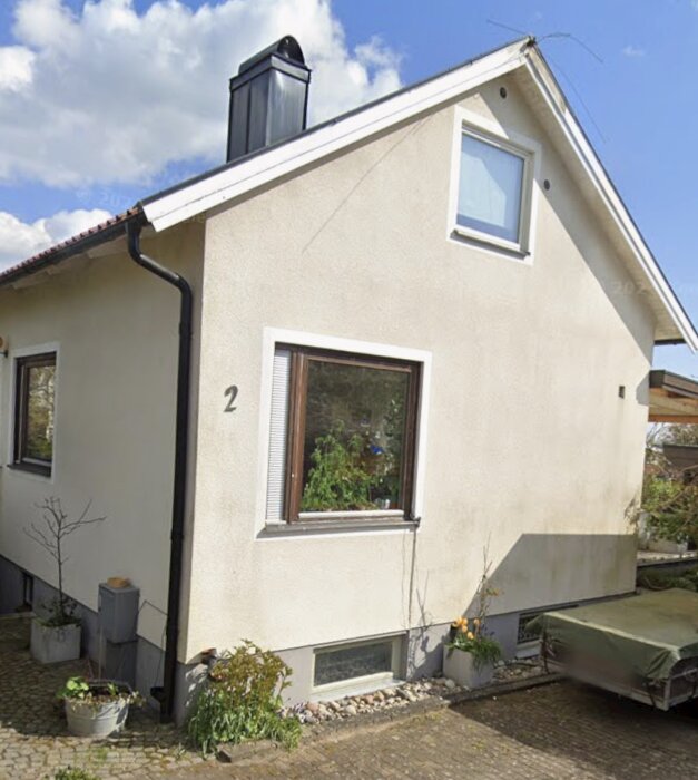 Färgat putsat litet hus med två fönster, en skorsten och ett enkelt tak. Omgiven av trädgård med växter och en släpvagn.