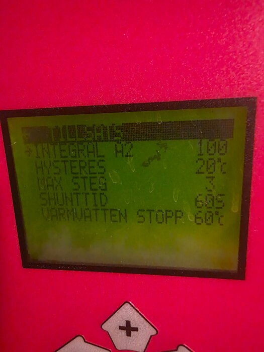Närbild av en digital display på en värmepump med inställningar för integralfrekvens, hysteres och varmvattenstopp, mot röd bakgrund.