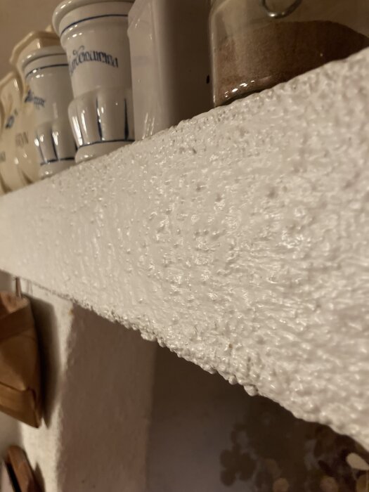 Rough surface of a kitchen hood with shiny white paint, surrounded by ceramic jars, questioning if the paint should be removed before refinishing.