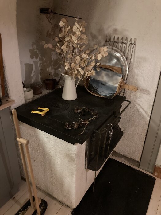 Gammal spis med dekoration ovanpå, inklusive en vit kanna med torkade växter, metallföremål och tavla, vid rustik vitmålad vägg.