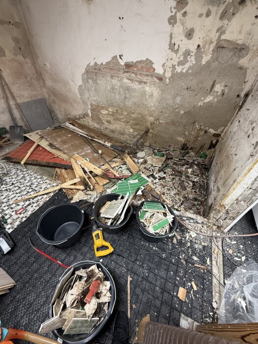Nedriven källardusch med synligt betonggolv, trasiga kakelplattor, träspån och verktyg som en skruvmejsel och hinkar med byggavfall.