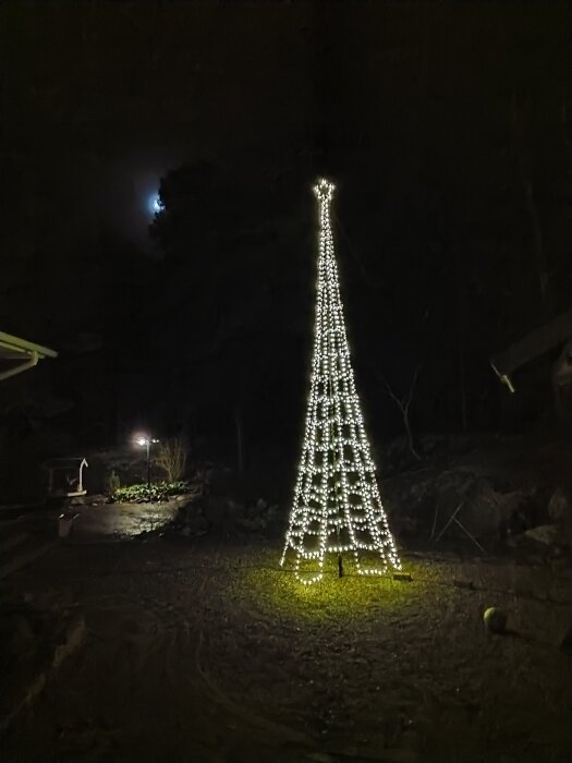 Konstgjort träd av ljusslingor lyser upp en mörk utomhusmiljö, med en synlig måne i bakgrunden.