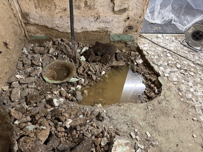 Riven källargrund där betong och stenkross har brutits upp, blottar en vattenpöl och en gammal golvbrunn i ett byggprojekt.