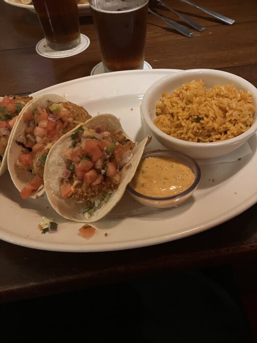 Shrimptacos med salsa och koriander, serverade med en skål ris och en dipsås på en tallrik, samt ett glas öl i bakgrunden.