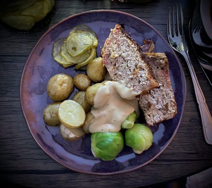 Köttfärslimpa med stekfläsk, brysselkål, nyskördade ugnsstekta potatisar, inlagd gurka och gräddsås med äppelgelé på en tallrik.