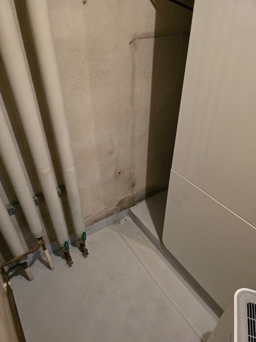 Betongvägg och golv i pannrum med synliga rör och fuktfläckar på väggen, troligen orsakade av avloppsläckage.