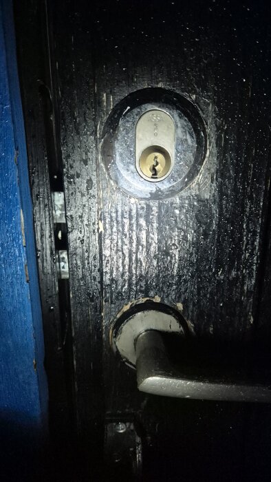 Närbild på ett lås och dörrhandtag på en mörk dörr med synlig glipa mellan dörr och karm.
