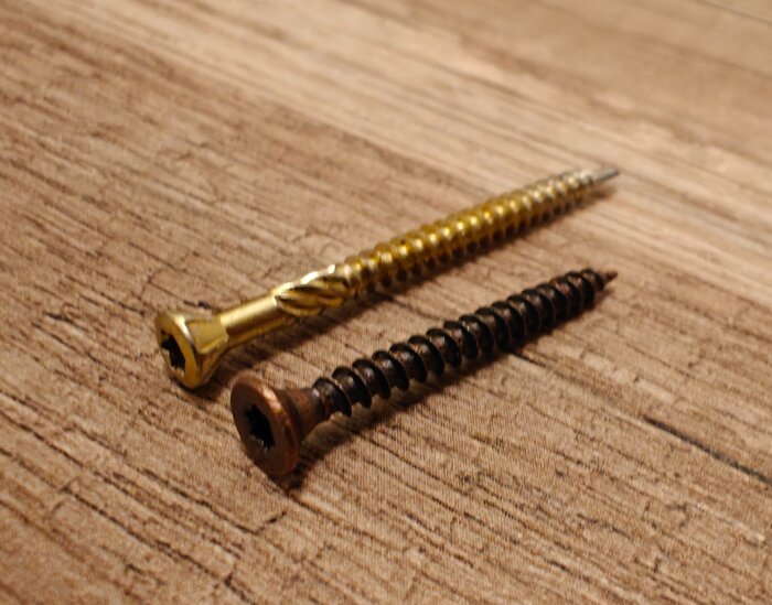 Två skruvar på ett trägolv, en guldig och en mörkare bronstonad, visas jämte varandra för att illustrera skillnaden i färg och design.