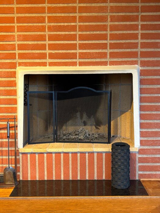 Öppen spis från 1963 med tegelvägg och svart gnistskydd, omgiven av verktyg och prydnad; planeras moderniseras med puts och insats.