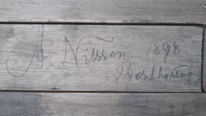 Handskriven text på en träplanka, daterad 1898, med ett namn och en ort.