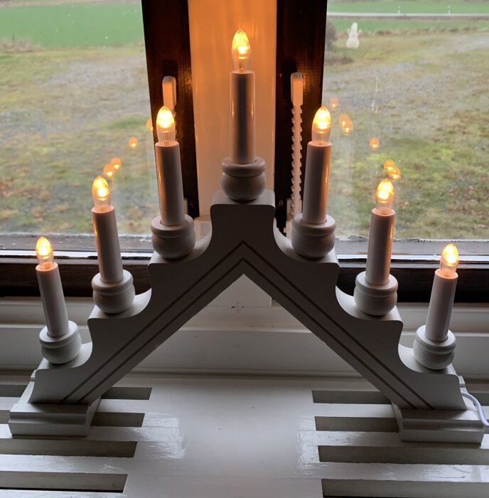Klassisk vit träljusstake med sju brinnande LED-lampor, placerad i ett fönster.