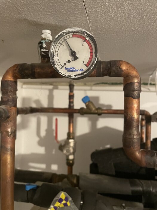 Tryckmätare monterad på rostigt rörledningssystem i källare, potentiellt igenkloggade slangar synliga i bakgrunden.