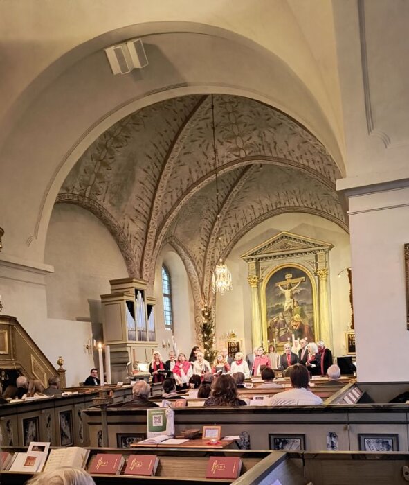 En kyrka med präst och kör framför altare med målning av Jesus på korset. Besökare sitter i bänkar med psalmböcker framför sig.