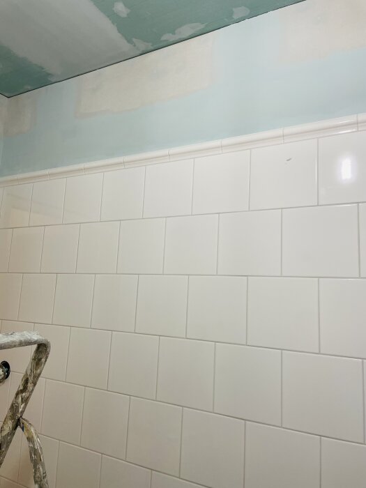 Kaklad vägg med vita plattor under ett delvis spacklat och målat tak i ett badrum. En ställning syns längst ner till vänster.