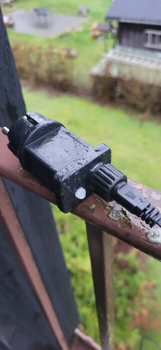 En svart elkontakt ligger på ett vått balkongräcke med en grönskande trädgård i bakgrunden.