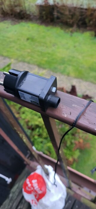 Elektrisk komponent med haneplugg och anslutningsmöjlighet, placerad på ett räcke med gräsmatta i bakgrunden.