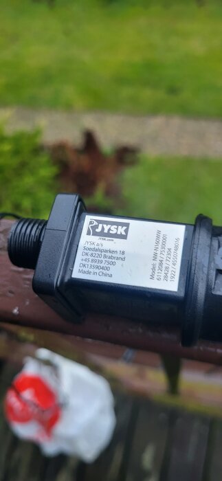 Närbild på en Jysk produktetikett med kontaktuppgifter. Produkten är svart och avlång, med bakgrund av en grönskande trädgård.