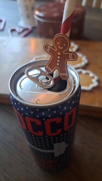 Ett sugrör med pepparkaksfigur sticker upp ur en öppnad läskburk, med pepparkakor i bakgrunden på ett bord.